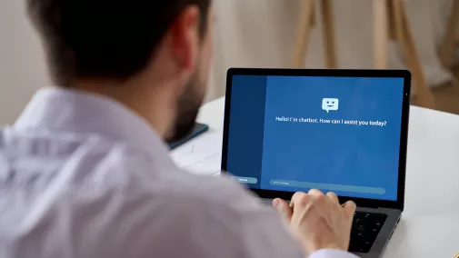 Foto von der Rückseite eines Mannes, der auf einen Laptop blickt. Auf dem Monitor sieht man einen Chatbot, den der Mann bedient.
